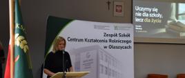 Zdjęcie przedstawia oficjalną uroczystość w Zespole Szkół Centrum Kształcenia Rolniczego w Oleszycach, podczas której kobieta przemawia przy mównicy na tle pamiątkowego baneru. W tle widoczny jest ekran z cytatem Seneki na temat nauki dla życia, a na pierwszym planie znajduje się fragment szkolnego sztandaru.