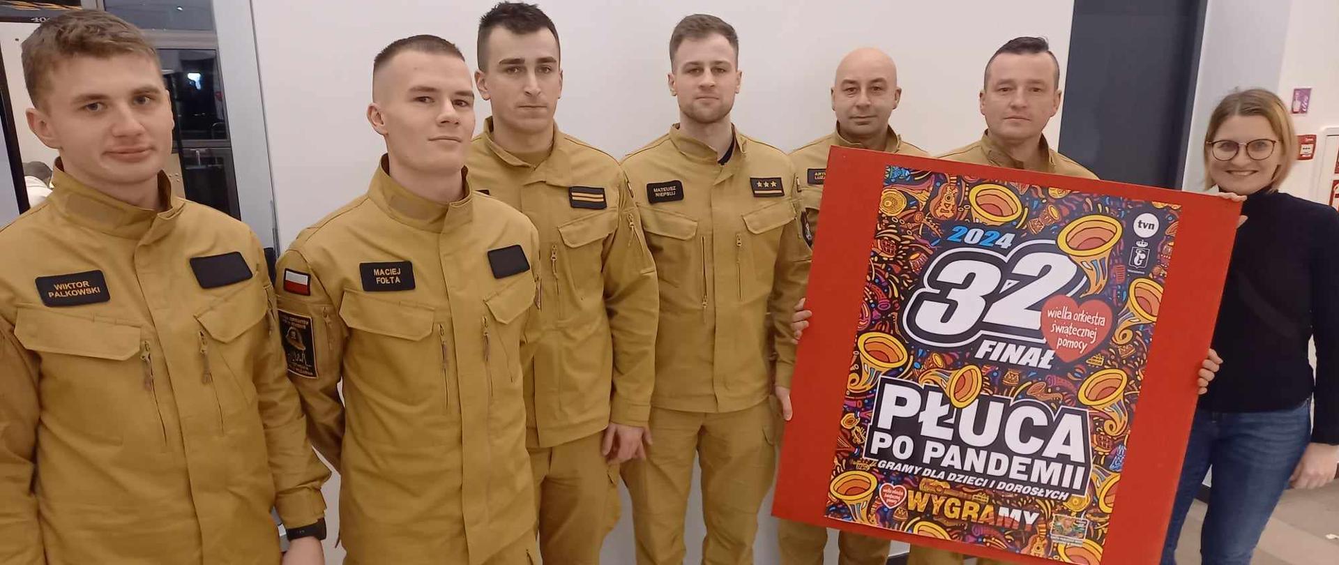 Pamiątkowe zdjęcie uczestników 32. Finału WOŚP (kadry i słuchaczy) jedna z osób trzyma baner z hasłem wydarzenia