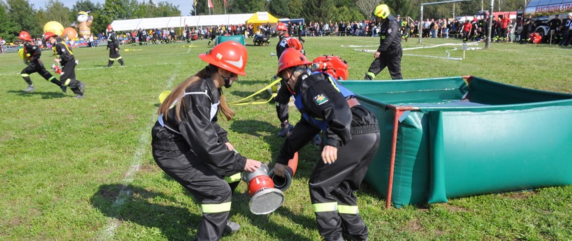 Kobieca drużyna pożarnicza w czarnych mundurach z czerwonymi hełmami na głowach podczas łączenia linii ssawnej