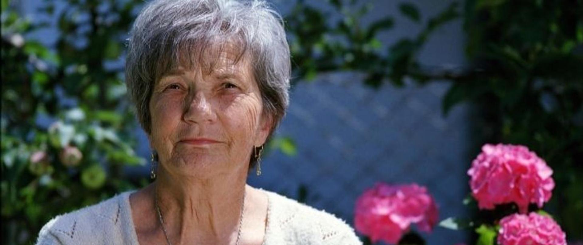 Na pierwszym planie starsza kobieta. Na drugim planie jabłoń, pelargonia i ogrodzenie z siatki