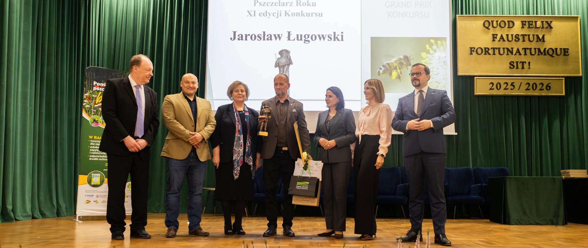Zdjęcie przedstawia 7 elegancko ubranych kobiet i mężczyzn na scenie. Na środku jest mężczyzna ze statuetką zwycięzcy