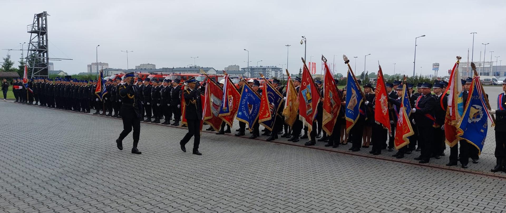 Na zdjęciu widoczne poczty sztandarowe jednostek OSP. Widzimy również 2 osoby oddające honor sztandarom. Apel odbywa się na terenie JRG 2, sztandary w momencie oddawania honorów są opuszczone do połowy.