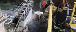 Zdjęcie przedstawia strażaków dogaszających pożar izolacji zbiornika. Znajdują się oni na platformie w górnej części zbiornika.