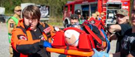 Ratownicy medyczni transportujący poszkodowanego na noszach, w tle samochód gaśniczy i inni uczestnicy ćwiczeń.