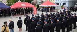 Strażacy stoją w pododdziałach, w tle trybuna honorowa oraz zaproszeni goście pod namiotem