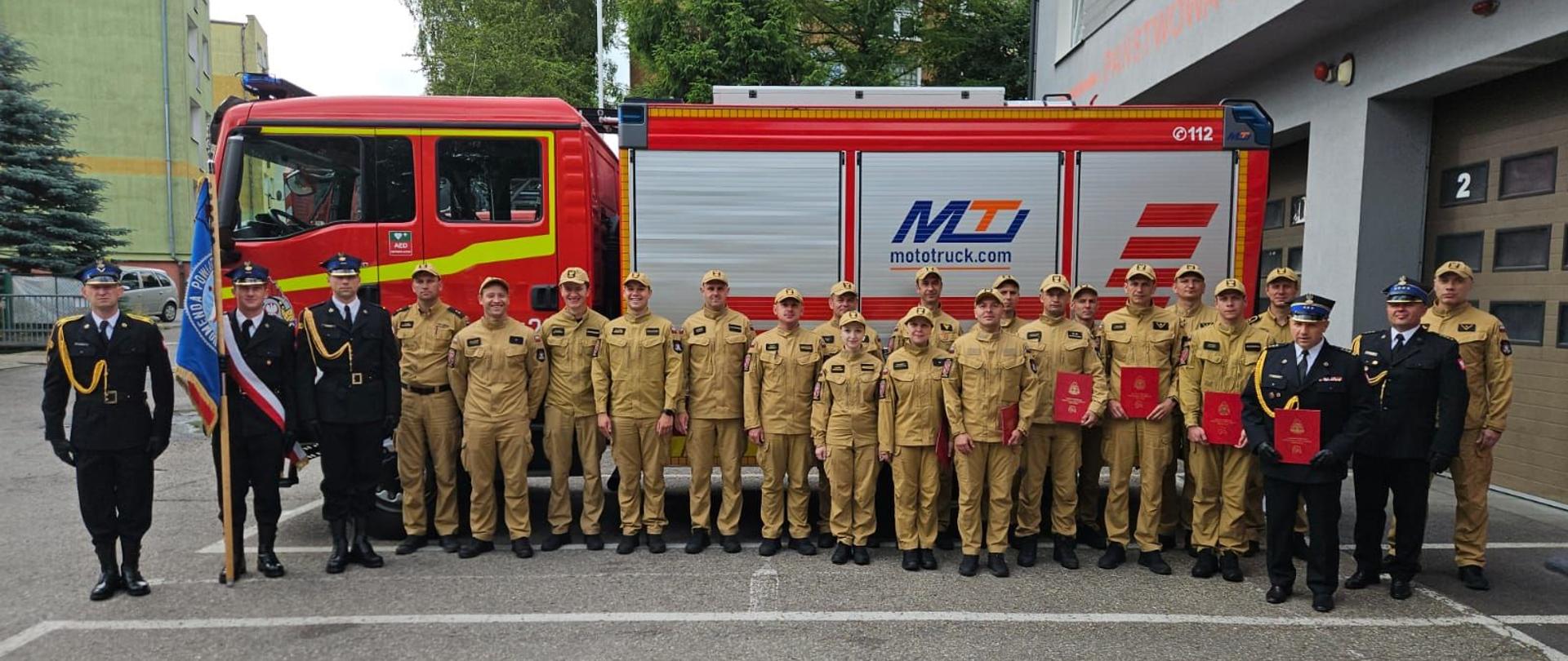 Na zdjęciu grupa strażaków. Z lewej trzech w mundurach galowych ze sztandarem. reszta w jasnych mundurach. Z prawej strony dwóch strażaków w galowych mundurach. W tle duży samochód strażacki.