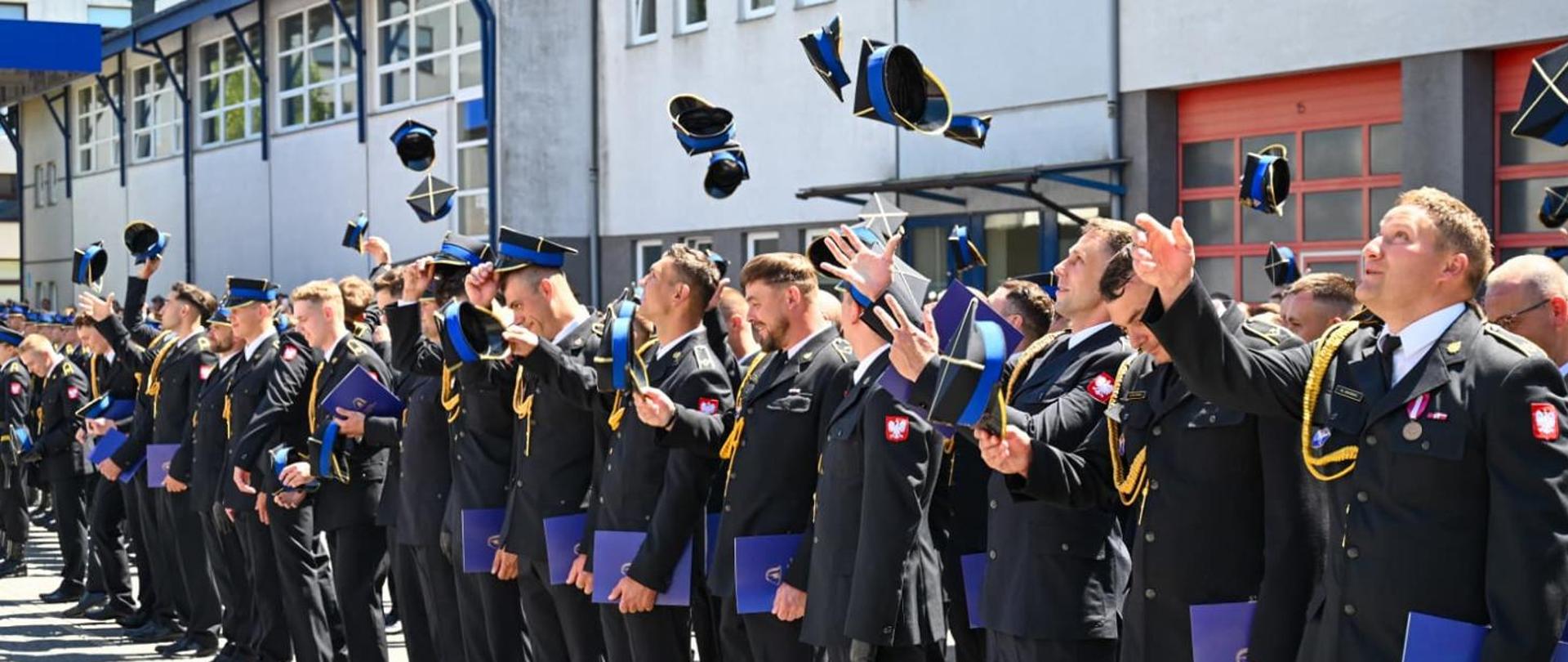 Na zdjęciu widać uroczystość zakończenia szkolenia lub promocji grupy funkcjonariuszy. Uczestnicy, ubrani w galowe mundury z przypiętymi odznakami i sznurami reprezentacyjnymi, stoją w rzędzie przed budynkiem. W kulminacyjnym momencie ceremonii podrzucają swoje czapki do góry
