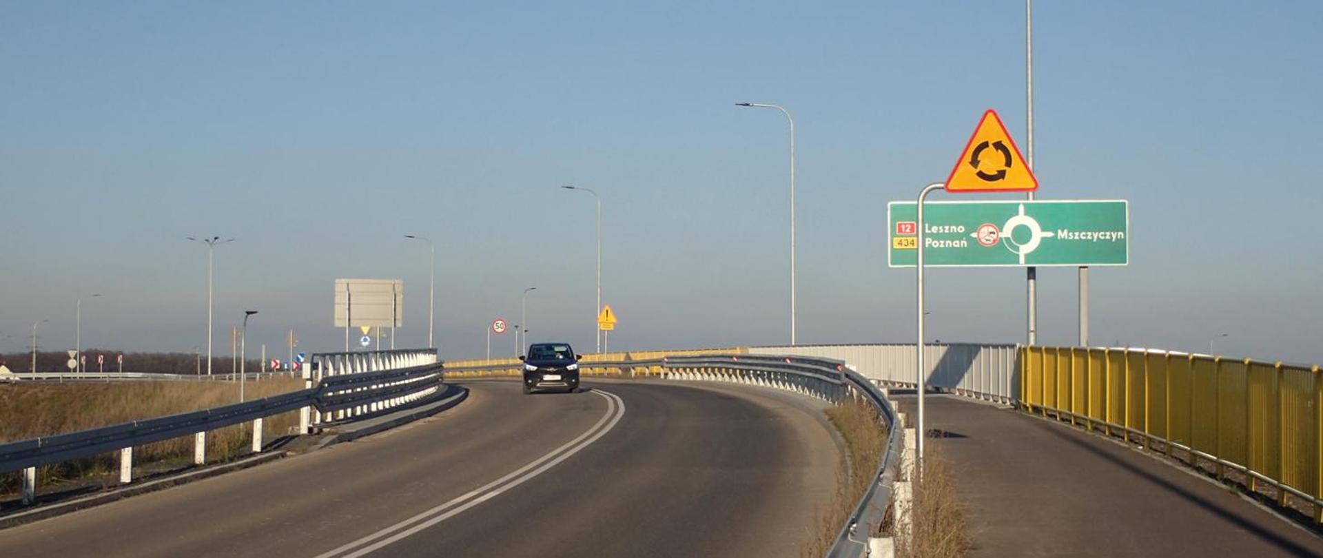 Droga na wiadukcie z barierkami i żółtymi ekranami, znak ostrzegawczy o rondzie oraz zielona tablica kierunkowa wskazująca Leszno, Poznań i Mszczyczyn. Na jezdni jedzie samochód, w tle widoczne latarnie i niebo bezchmurne.