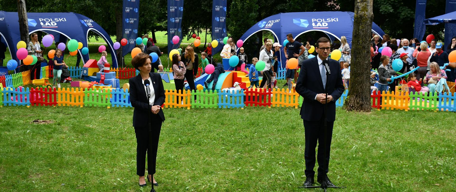 Na zdjęciu znajdują się dwie elegancko ubrane osoby - kobieta i mężczyzna. Stoją oni na trawie, przed mikrofonami i są w trakcie wystąpienia publicznego. W tle znajduje się tymczasowy plac zabaw dla dzieci, z kolorowym, niskim ogrodzeniem. Na dalszym planie znajdują się granatowe namioty oraz flagi z napisami "Polski Ład".