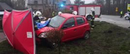 Strażacy ratownicy uwalniają przy pomocy sprzętu hydraulicznego uwiezionego pasażera samochodu osobowego. Samochód znajduje się poza jezdnią na psie zieleni. Policja kieruje ruchem drogowym