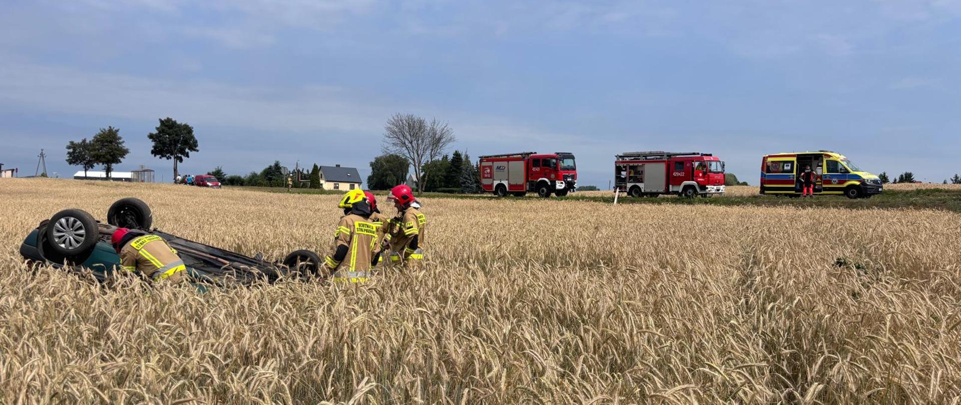 Na zdjęciu widać samochód osobowy w pozycji na dachu, który wypadł z drogi i zatrzymał się na przydrożnym polu w rosnącym zbożu. Przy samochodzie są strażacy a w tle na drodze stoją pojazdy służb ratunkowych.