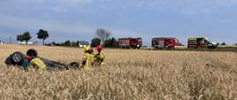 Na zdjęciu widać samochód osobowy w pozycji na dachu, który wypadł z drogi i zatrzymał się na przydrożnym polu w rosnącym zbożu. Przy samochodzie są strażacy a w tle na drodze stoją pojazdy służb ratunkowych.