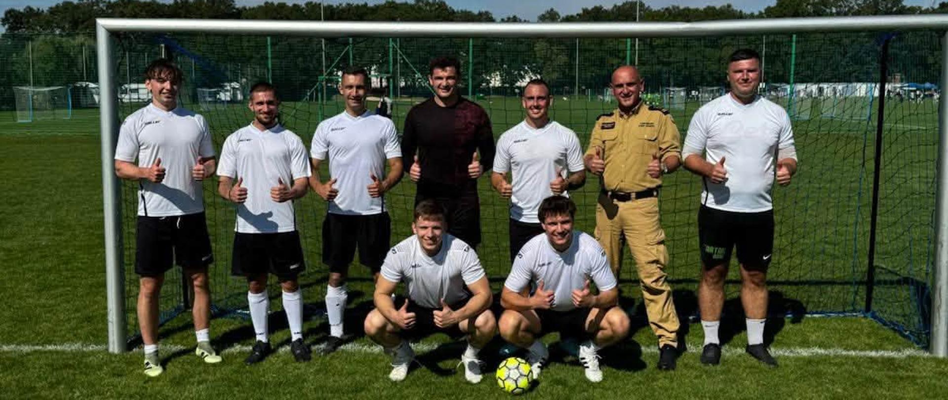 Reprezentacja KM PSP Wrocław piłkarzy wraz z Komendantem Miejskim PSP we Wrocławiu pozują do wspólnego zdjęcia w bramce na zielonym boisku piłkarskim w słoneczny dzień