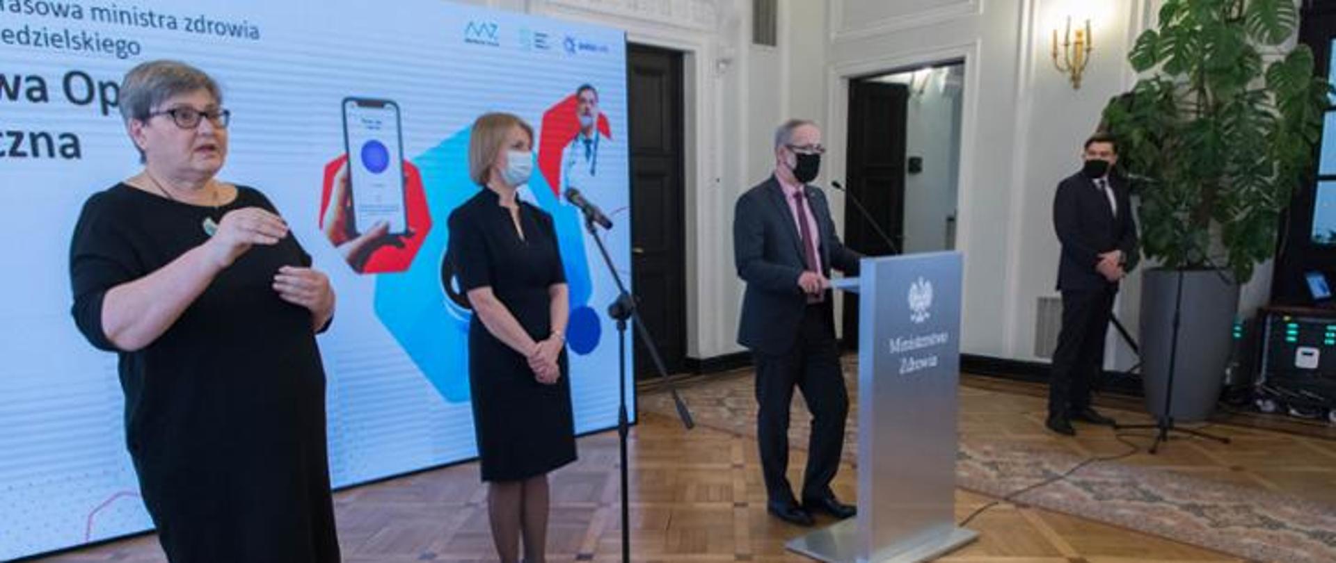 konferencja prasowa w pomieszczeniu, 4 osoby stoją, do mikrofonu mówi minister zdrowia Adam Niedzielski, w tle banner z aplikacją programu DOM