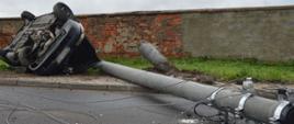 Kolizja w Trzeboszu. Auto osobowe uderzyło w słup energetyczny. Samochód leży na lewym boku. Obok na jezdni znajduje się złamany betonowy słup oraz zerwane przewody energetyczne. W tle murowane ogrodzenie gospodarstwa.