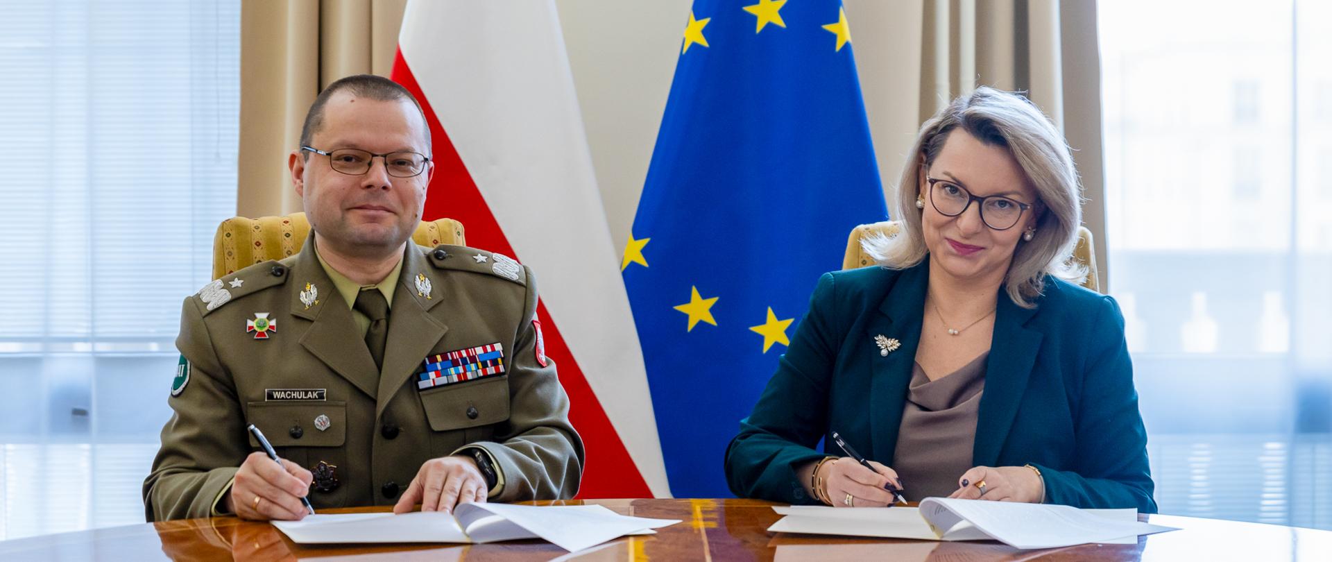 Dyrektor Generalna Ministerstwa Finansów Marta Niżałowska-Pactwa oraz Rektor - Komendant WAT gen. bryg. Przemysław Wachulak, przy biurku podpisują dokumenty. W tle flagi Polski i UE