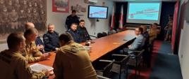 Na zdjęciu widać grupę mężczyzn siedzących wokół długiego stołu w sali konferencyjnej. Większość z nich ma na sobie mundury, w tym granatowe mundury policyjne i beżowe strażackie. Wszyscy siedzą, wpatrując się w prezentację wyświetlaną na dużym ekranie na końcu sali.