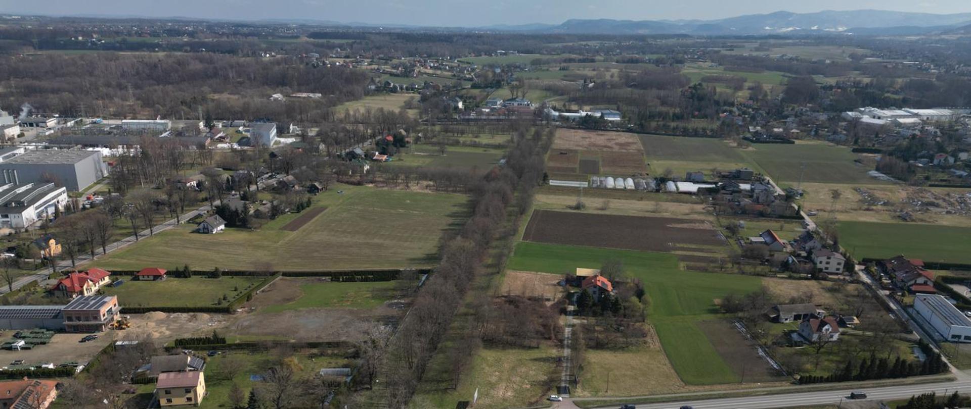 Obraz przedstawia widok z lotu ptaka na wiejski krajobraz z polami, domami i drzewami. Widać kilka zielonych pól, brązową zaoraną ziemię oraz rozrzucone domy z czerwonymi dachami. Na dole obrazu widoczna jest droga, a po lewej stronie znajdują się budynki przemysłowe. W tle widać pasmo wzgórz.