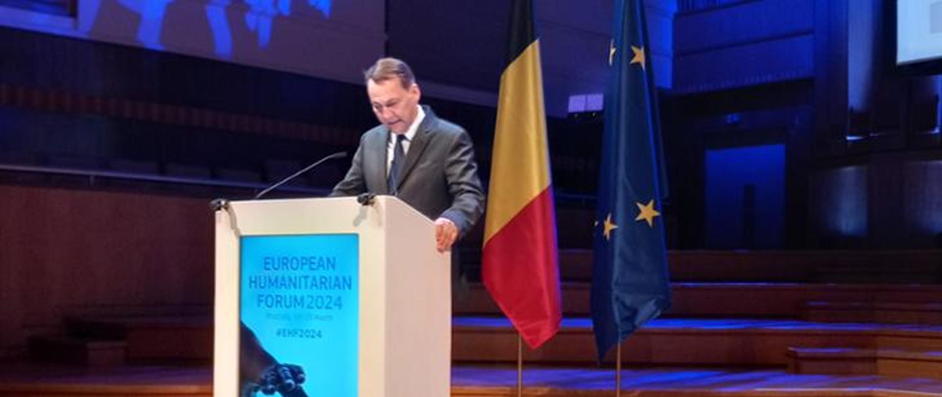 Mężczyzna w garniturze przemawia z podium z flagami Belgii i Unii Europejskiej w tle.