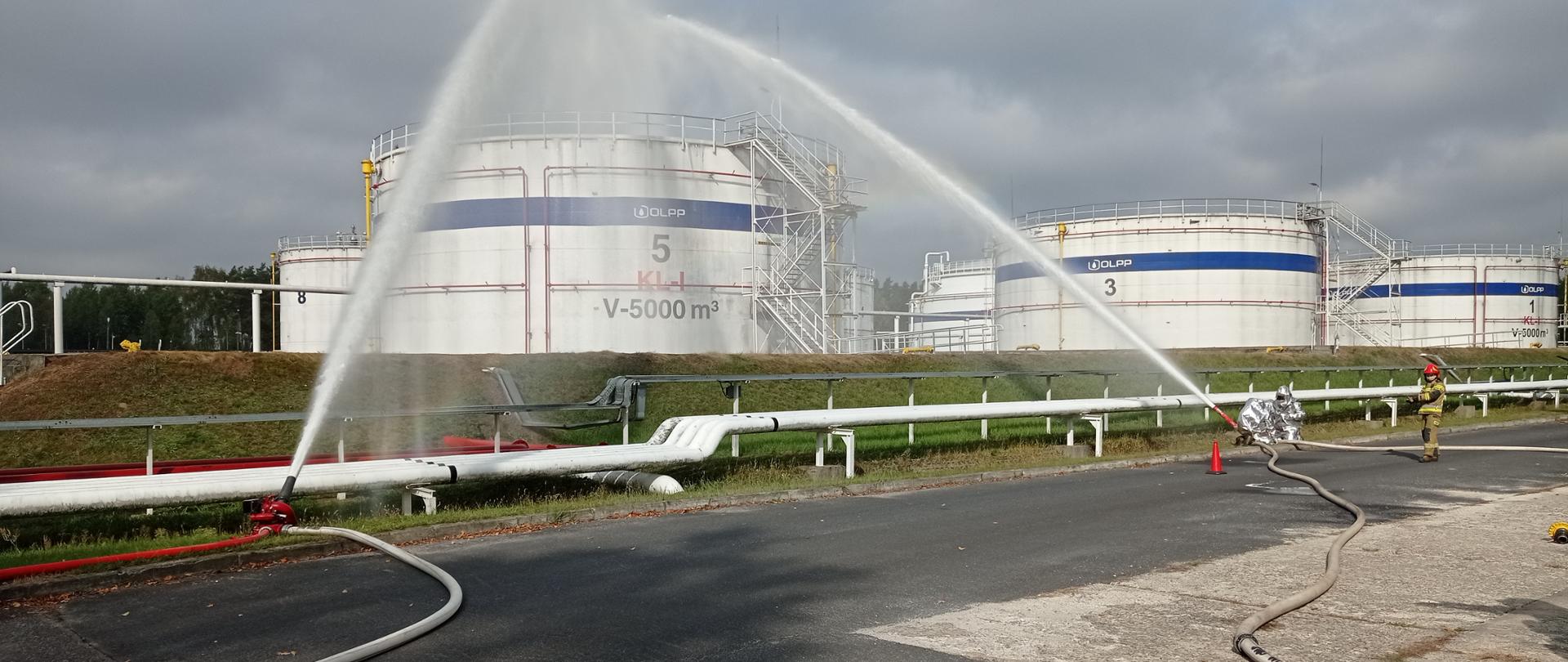 Na zdjęciu widoczne są duże zbiorniki wypełnione benzyną. Przed zbiornikami na asfaltowej drodze ustawione są działka wodno-pianowe podające wodę na zbiorniki. Nieopodal działek stoją strażacy w srebrnych ubraniach żaroodpornych. Przed strefą promieniowania cieplnego widoczny jest strażak w piaskowym mundurze oraz w czerwonym hełmie na głowie. Tło zdjęcia obrazuje zachmurzone niebo.
