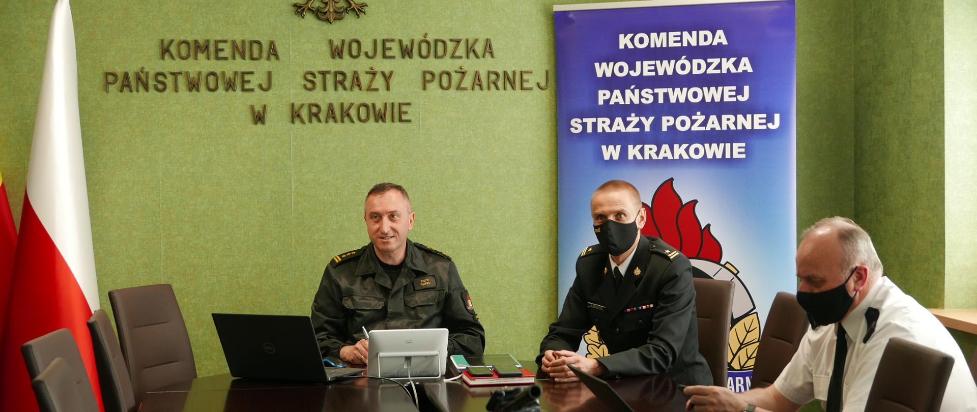 Komendant Wojewódzki PSP wraz z Zastępcą i Komendantem Miejskim PSP w trakcie trwania wideokonferencji. Po prawej stronie baner Komendy Wojewódzkiej, po lewej flagi.