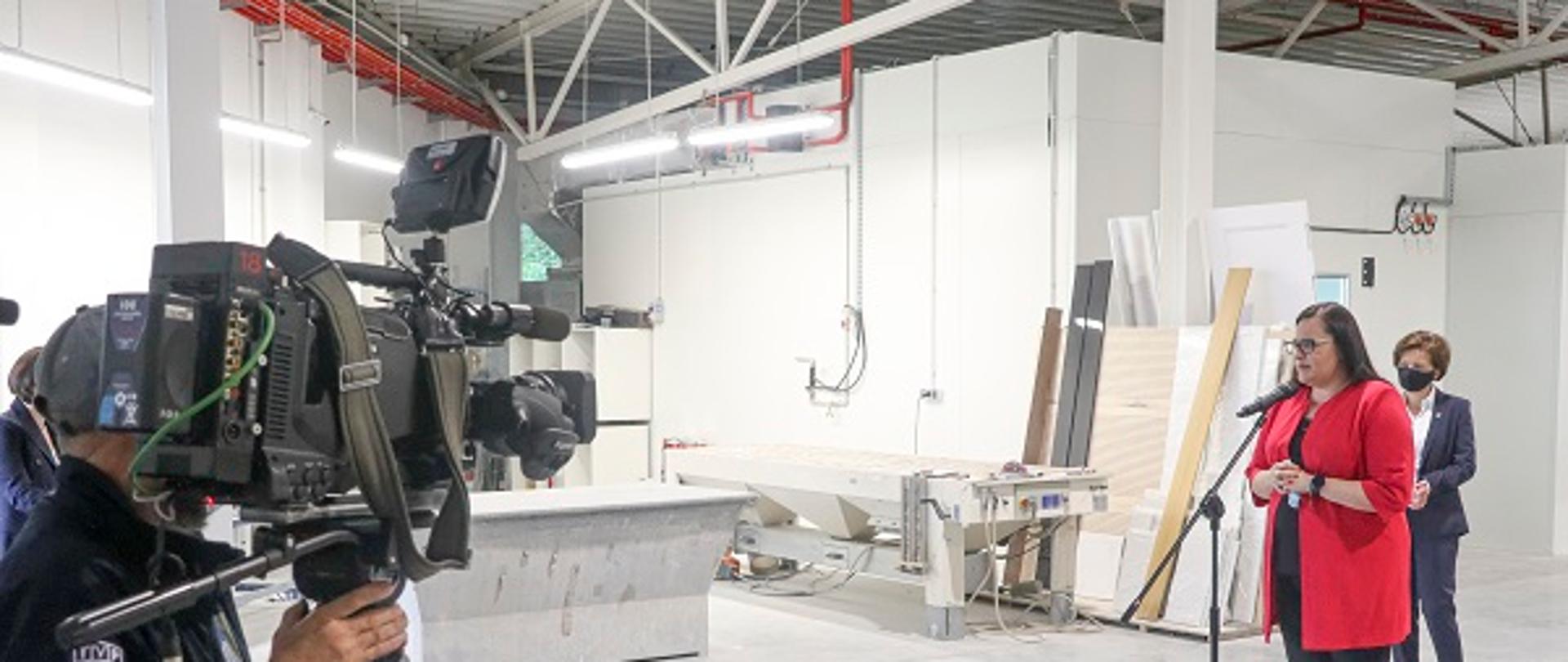 Minister Małgorzata Jarosińska-Jedynak przemawia przed mikrofonem w hali produkcyjnej firmy. Za jej plecami stoi minister Marlena Maląg. Z przodu widoczny operator z kamerą.