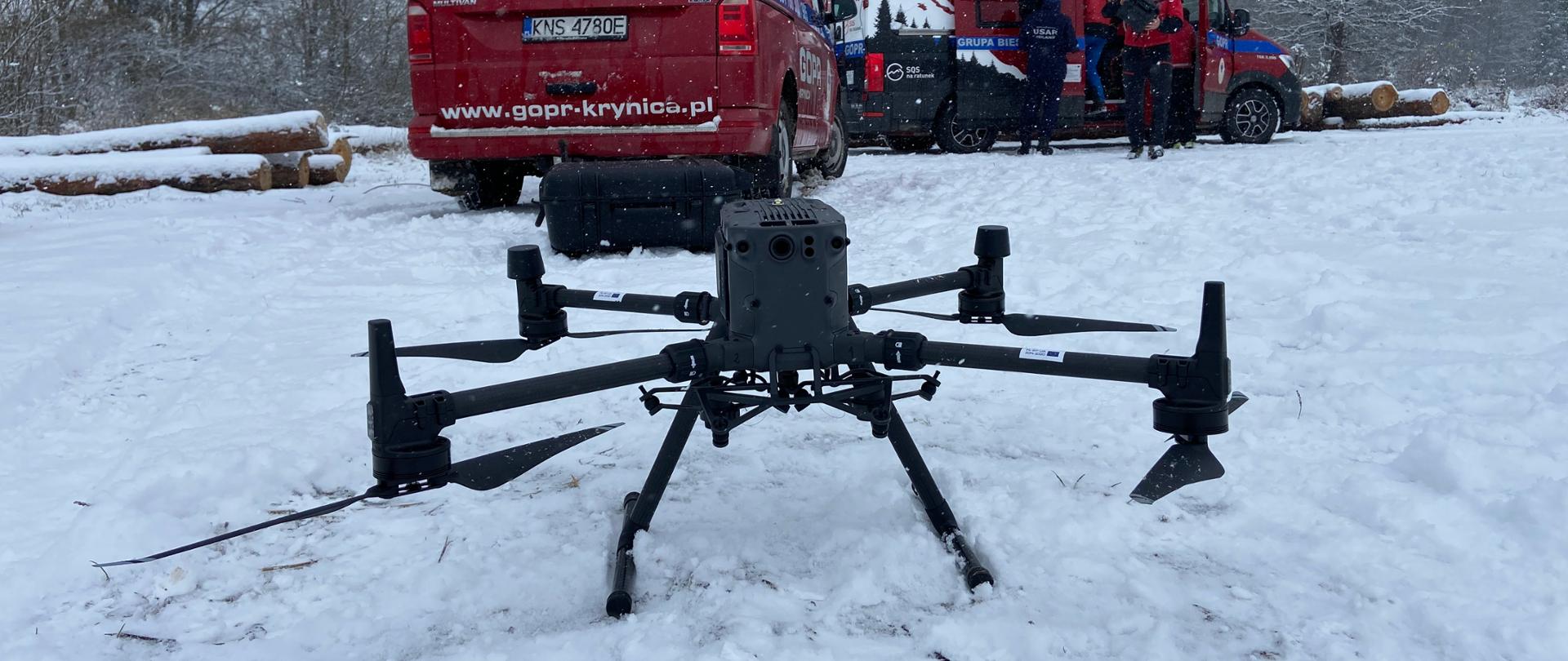 Na pokrytej śniegiem łące na pierwszym planie ustawiony jest dron (quadrocopter) tło stanowią samochody operacyjne (busy) Górskiego Ochotniczego Pogotowia Ratunkowego