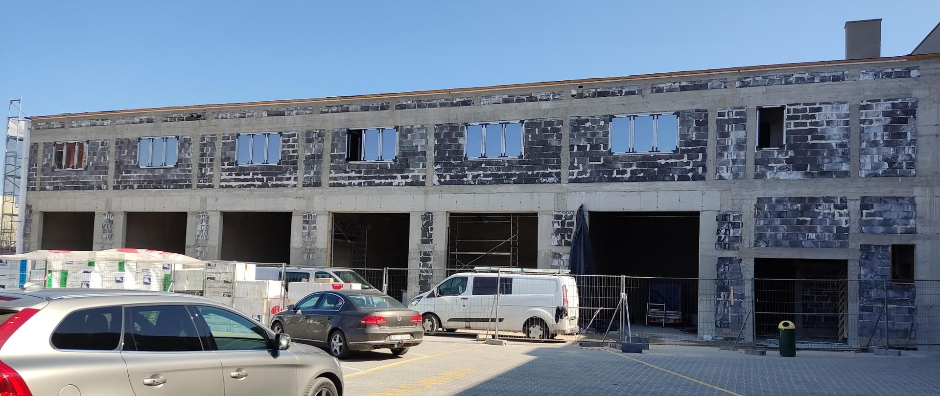 Na zdjęciu budowany obiekt garażowy. Widać również materiały budowlane oraz zaparkowane przed budynkiem na parkingu samochody.