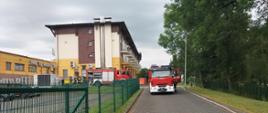 W tle budynek sanatorium, na którym prowadzone były ćwiczenia. na 1 planie po wykostkowanym wjeździe po prawej stronie rozciągnięte weże pożarnicze, na wjeździe samochód pożarniczy oraz w tle przed budykiem, obok wozu strażackiego stoją strażacy,