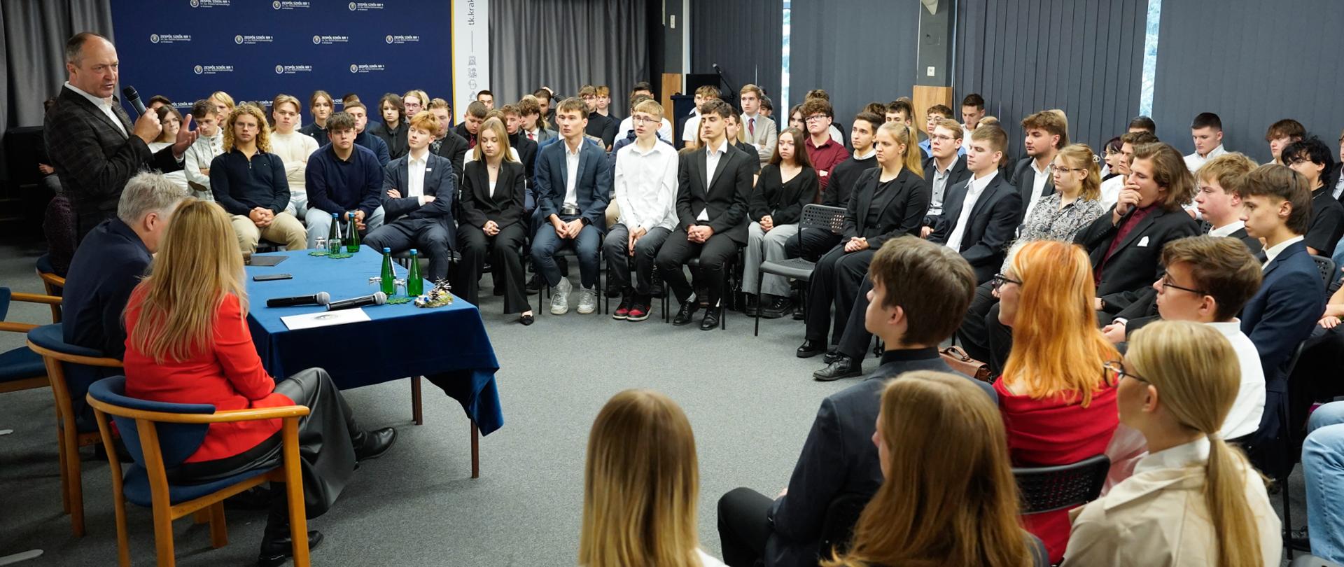 Zdjęcie przedstawia panel dyskusyjny lub spotkanie w dużej sali konferencyjnej. Na pierwszym planie, z tyłu, siedzą dwie osoby dorosłe przy niebieskim stole, a na pierwszym planie, z przodu, widoczne są tyły głów innych osób dorosłych. Po lewej stronie stoi mężczyzna w garniturze przemawiający do publiczności, trzymając mikrofon. Publiczność, składająca się głównie z młodzieży w strojach formalnych (część w garniturach i garsonkach), siedzi w rzędach. W tle widoczna jest ciemnoniebieska ściana z białymi napisami.