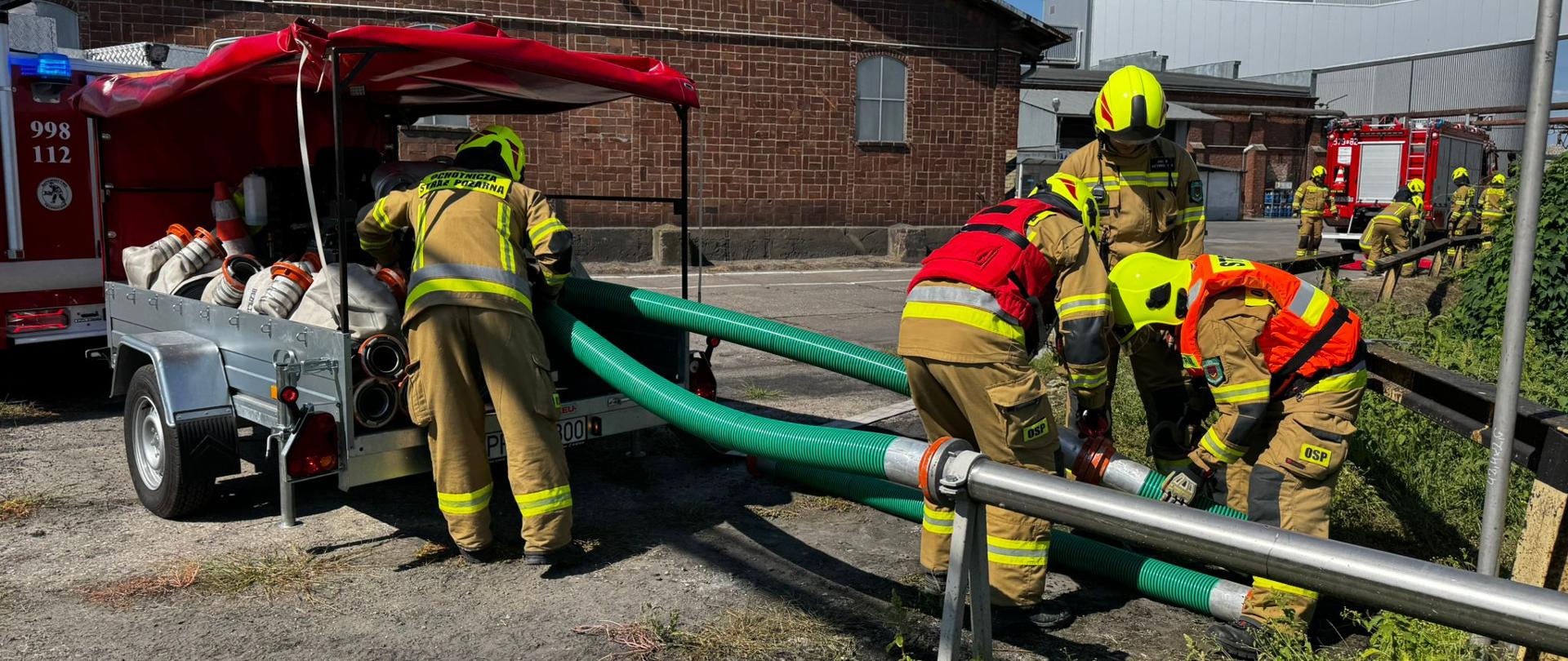 Strażacy ochotnicy podłączają zielone węże ssawne do pompy na przyczepie wężowej. W tle ceglany budynek zakładu, silos cukru oraz wozy strażackie. Na pierwszym planie intensywne działania związane z budową magistrali wodnej.