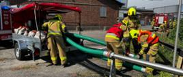 Strażacy ochotnicy podłączają zielone węże ssawne do pompy na przyczepie wężowej. W tle ceglany budynek zakładu, silos cukru oraz wozy strażackie. Na pierwszym planie intensywne działania związane z budową magistrali wodnej.