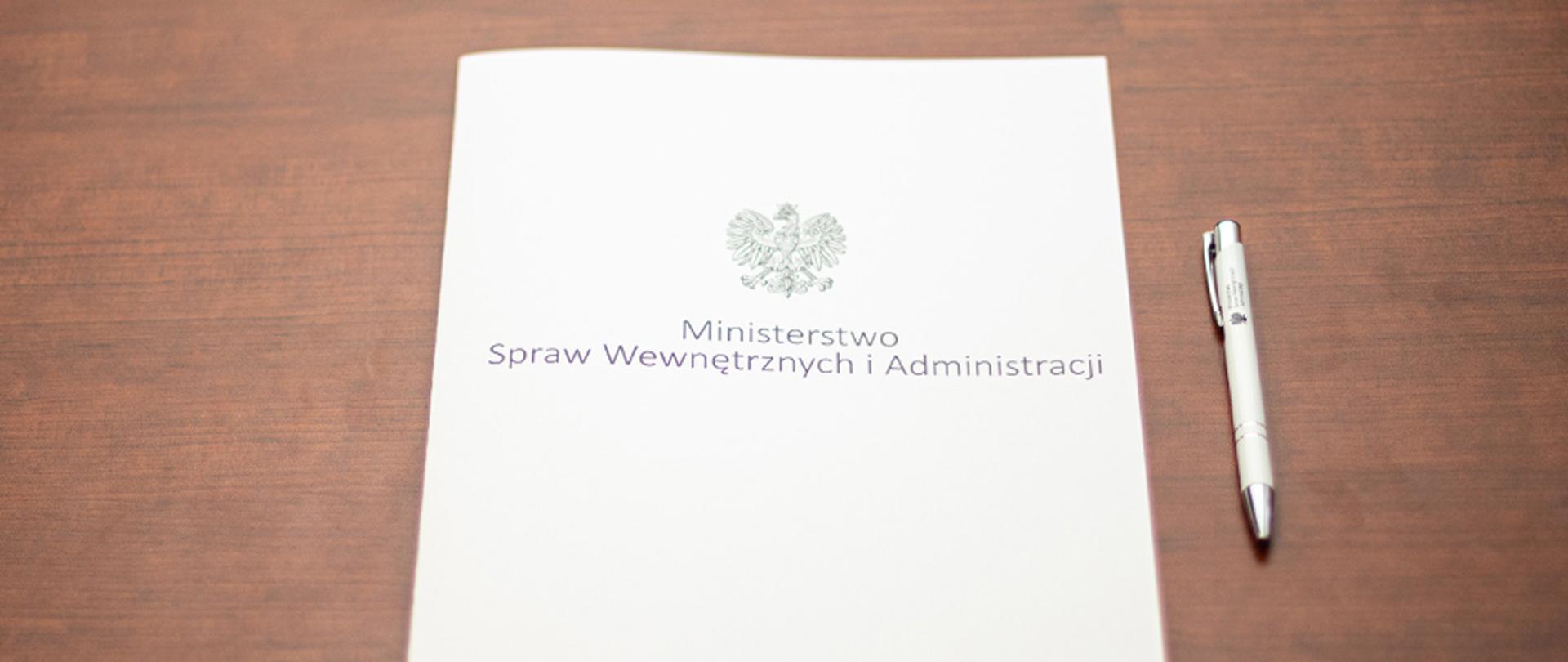 Na zdjęciu widać leżącą teczkę z napisem "Ministerstwo Spraw Wewnętrznych i Administracji" i orłem z godła narodowego i leżący obok długopis.