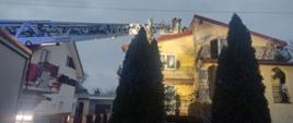 Strażacy na dachu budynku mieszkalnego prowadza działania gaśnicze. Jeden ze strażaków znajduje się w koszu drabiny mechanicznej