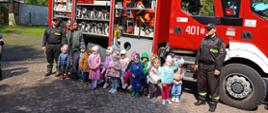 Zdjęcie przedszkolaków z 2 strażakami PSP na tle wozu strażackiego z otwartymi skrytkami ze sprzętem. 