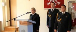 Na zdjęciu strażacy stoją w mundurach wyjściowych, jedna osoba przemawia za mównicą, na ścianie wisi godło Polski i Krzyż.