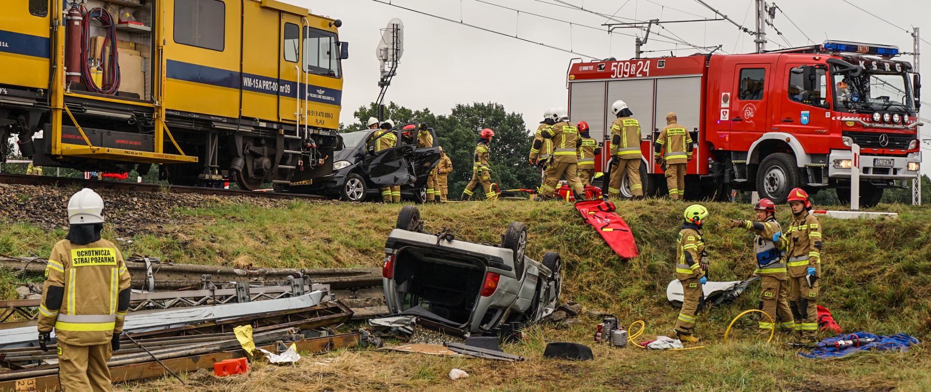 Na zdjęciu widać miejsce prowadzonych ćwiczeń. W centralnym punkcie samochód leżący na dachu u podnóża skarpy kolejowej z wyciętą klapą bagażnika. Na skarpie kolejowej lokomotywa PKP wraz z wrakiem samochodu osobowego, wokół którego ćwiczą strażacy w ubraniach bojowych. Obok wóz bojowy Ochotniczej Straży Pożarnej Rusinowice