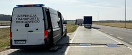 Samochód Inspekcji Transportu Drogowego zaparkowany przy drodze, obok rozstawionej przenośnej wagi do kontroli pojazdów. Na drugim planie kilka zatrzymanych do kontroli dostawczaków.