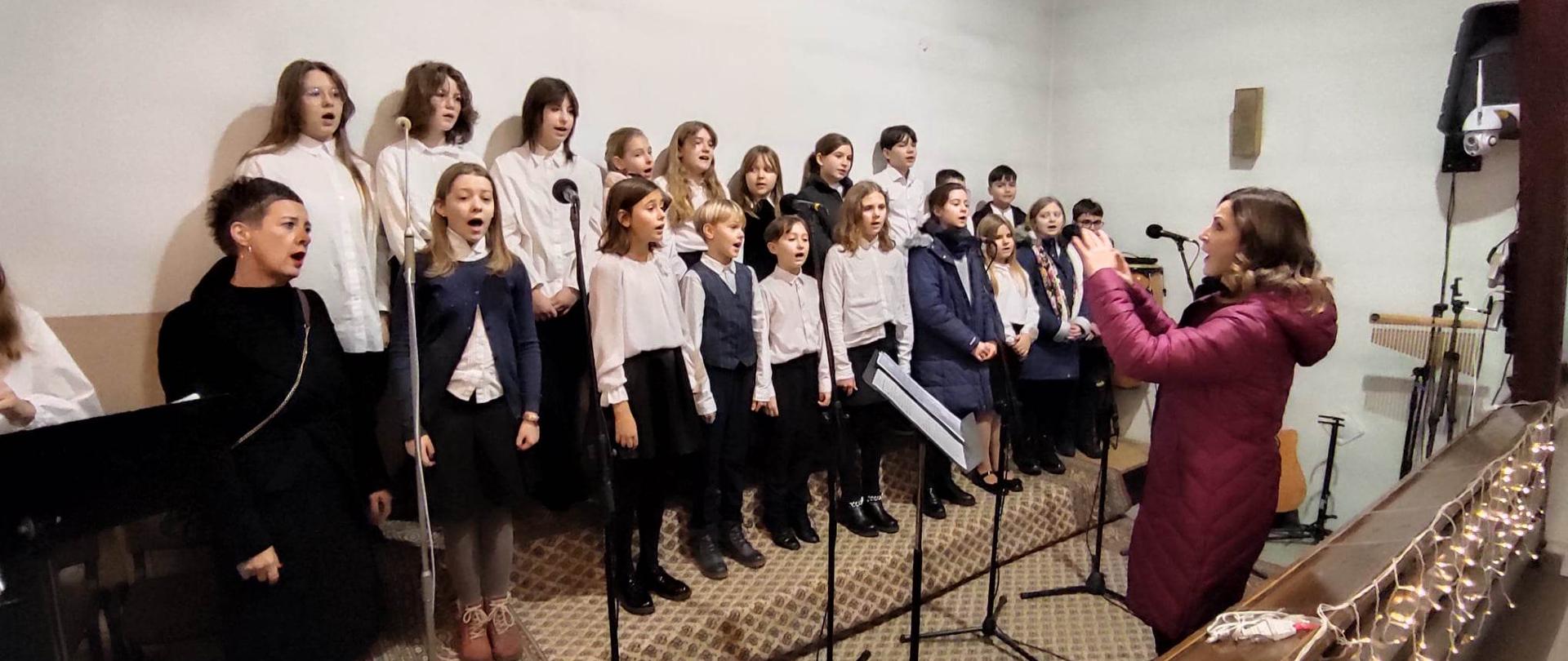 Zdjęcie chóru OSM I st. w Piszu na tle białej ściany kościoła. Dyryguje Agata Rożniatowska.