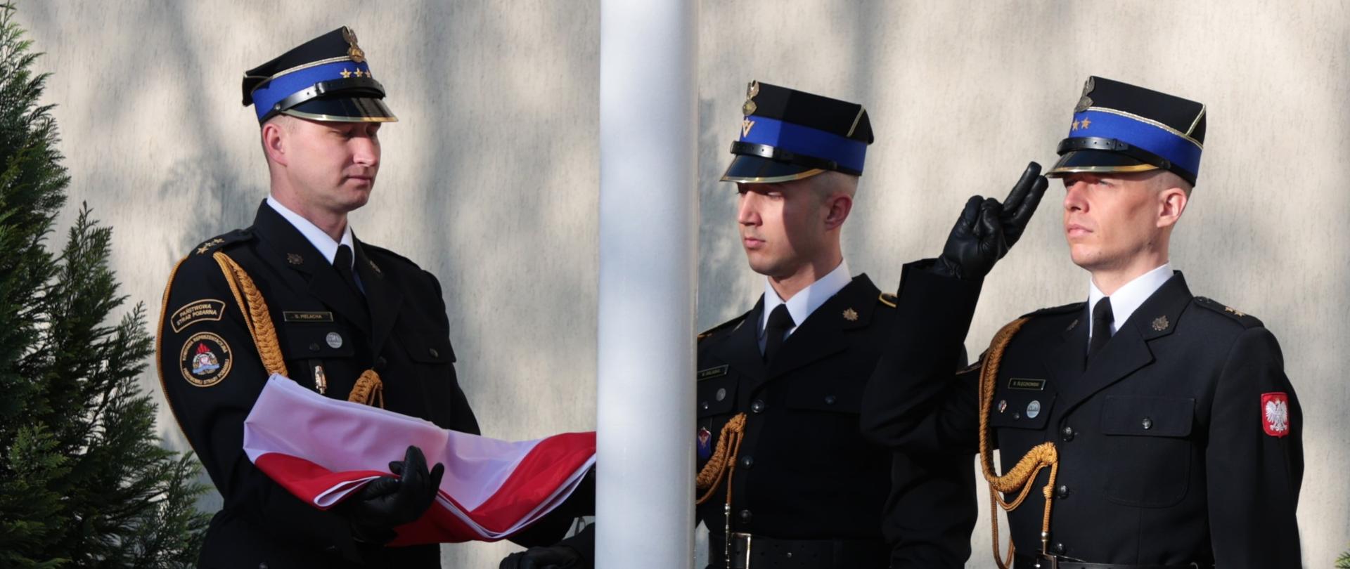 Poczet sztandarowy - 3 strażaków w mundurach galowych, strażak po lewej stronie trzyma na rękach złożoną flagę RP