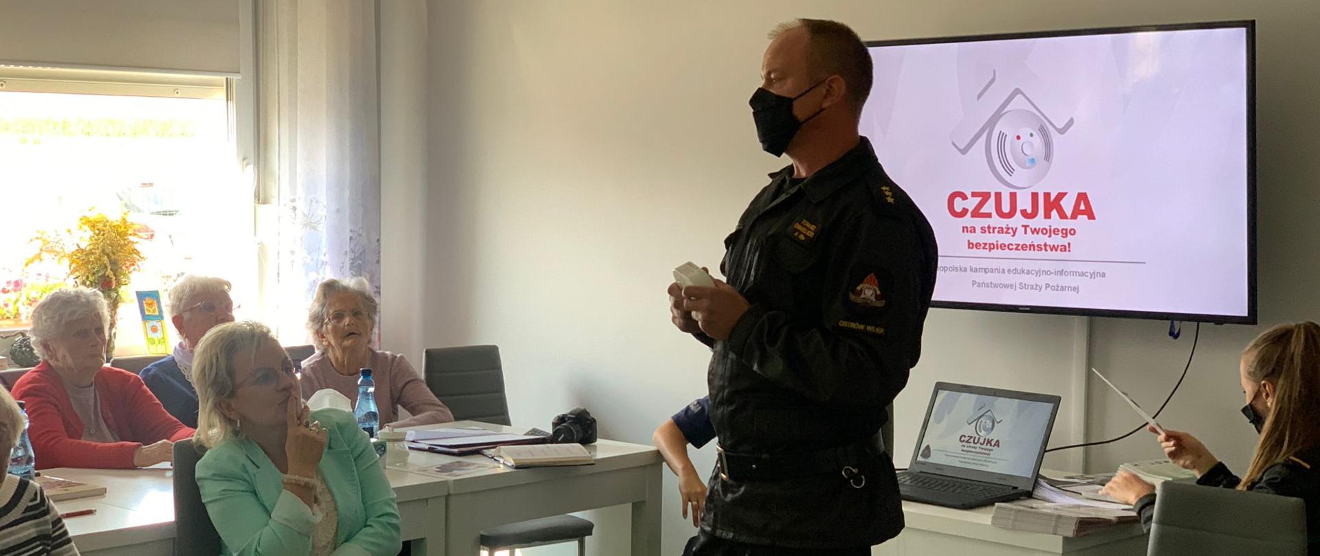 Funkcjonariusz trzyma w rękach czujkę dymu i tlenku węgla, opowiadając seniorom w sali wiejskiej o bezpieczeństwie pożarowym w domach.
