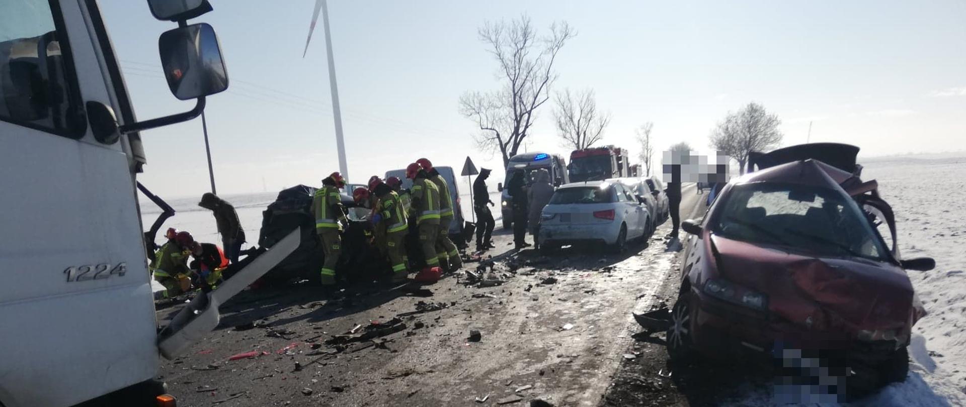 Na poboczu drogi stoi kompletnie rozbity pojazd osobowy. Po przeciwnej stronie jezdni widać drugi z rozbitych pojazdów, przy którym strażacy udzielają pomocy medycznej. Trwa ewakuacja osoby poszkodowanej. W kolumnie pojazdów rozbitych znajdują się kolejne trzy pojazdy, które zostały uszkodzone w wyniku wypadku. Droga całkowicie zablokowana. Pojazdy służb ratunkowych znajdują się na jednym pasie jezdni. 