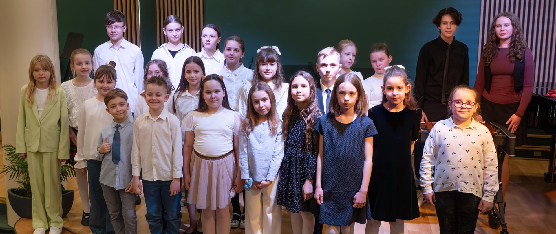 Grupa dzieci i młodzieży stoi na scenie w eleganckich strojach, prawdopodobnie po występie w szkole muzycznej. Na zielonej ścianie w tle widnieje logo z literami „PSM”. Dzieci mają różne stroje – niektóre ubrane są w klasyczne białe koszule i ciemne spodnie lub spódnice, inne w sukienki, a jedna dziewczynka ma jasnozielony garnitur. Większość ma poważne lub lekko uśmiechnięte wyrazy twarzy, a jedno z młodszych dzieci pokazuje kciuk w górę. W tle znajdują się elementy wystroju sali koncertowej, w tym fortepian i drewniane panele na ścianach.
