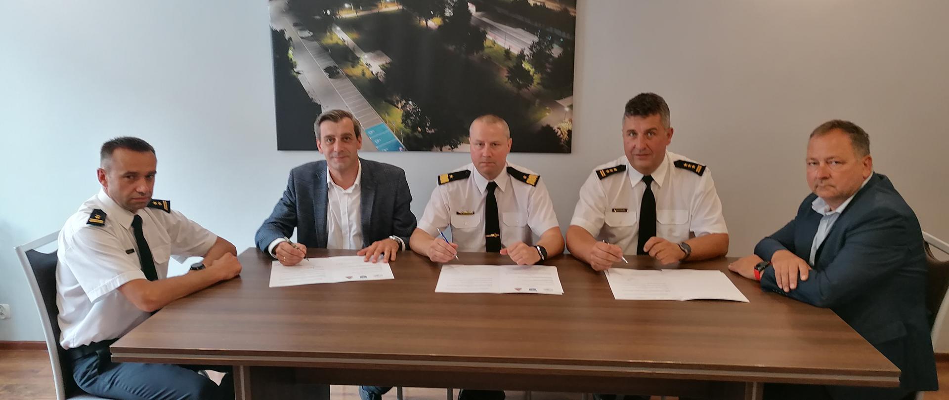 pięciu mężczyzn siedzi przy stole, przed nimi leżą teczki do podpisu 