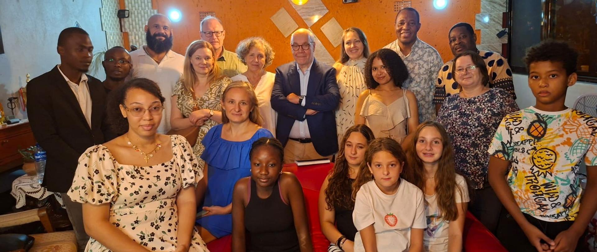 A group of 19 people gathered for a meeting of the Polish diaspora in Maputo; in the foreground, a group of children and young people sit on a red sofa and armchairs; a group of adults poses in the second row.