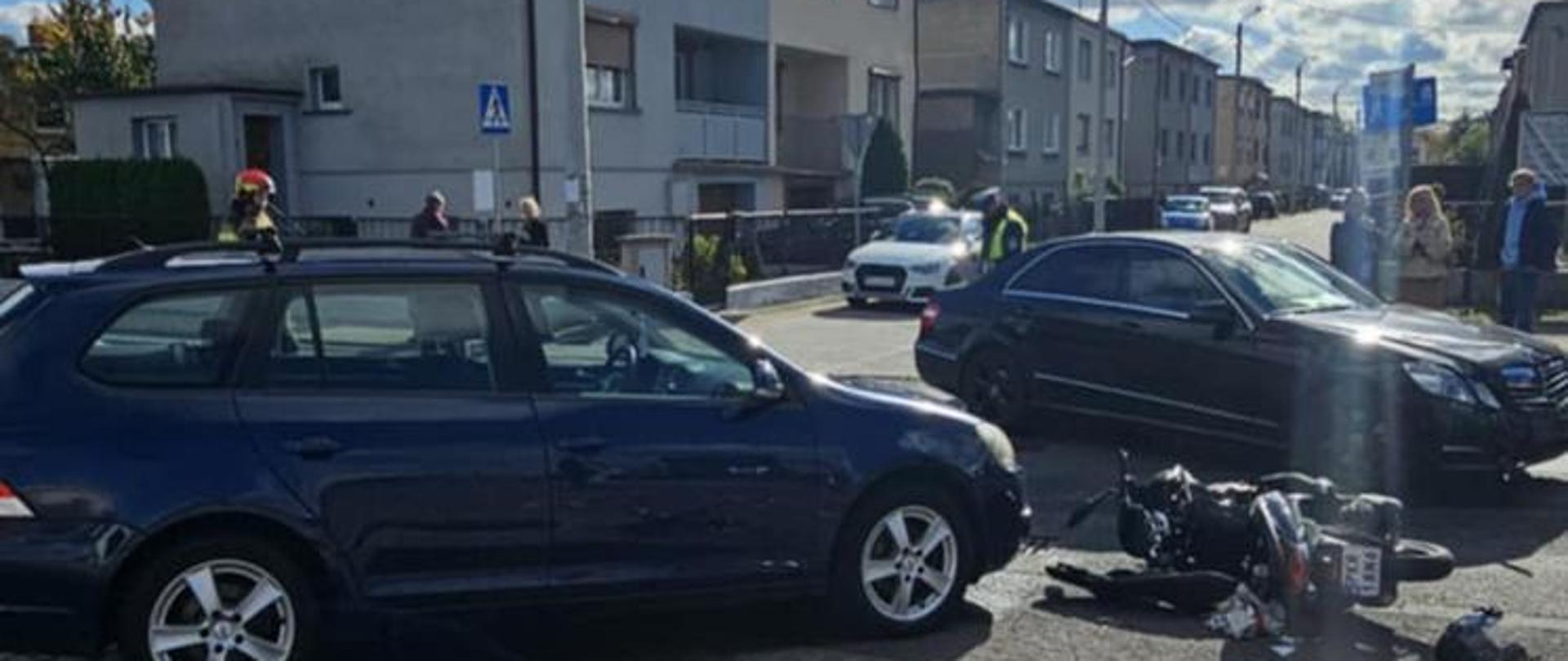 Na zdjęciu widać miejsce wypadku drogowego na miejskiej ulicy. Na pierwszym planie leży przewrócony motocykl oraz rozrzucone przedmioty, w tym plastikowe butelki i opakowania. Obok stoją dwa samochody osobowe – ciemny kombi i czarny sedan – które wyglądają na uczestników kolizji. W tle widać kilka osób, budynki mieszkalne i przejście dla pieszych. Pogoda jest słoneczna, na niebie widoczne są chmury.
