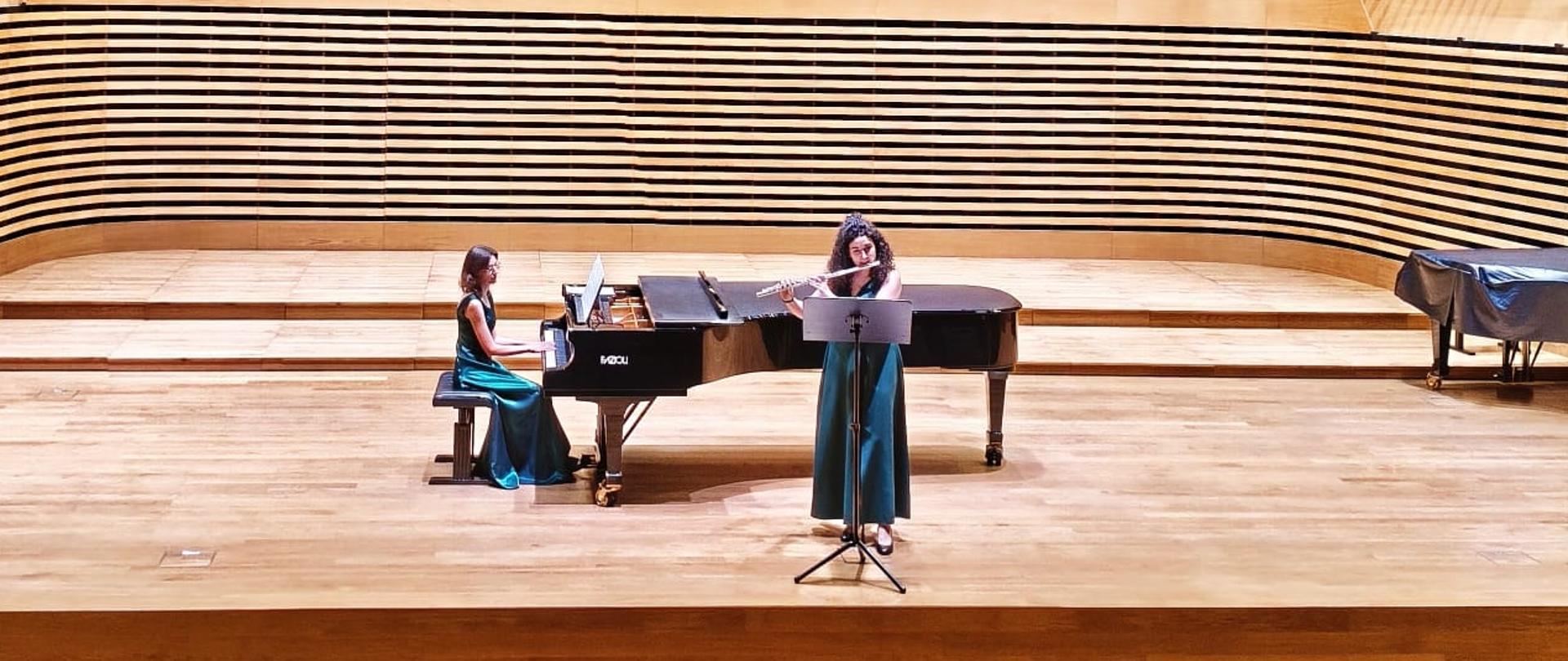 Dziewczyna gra na flecie, kobieta akompaniuje na fortepianie.