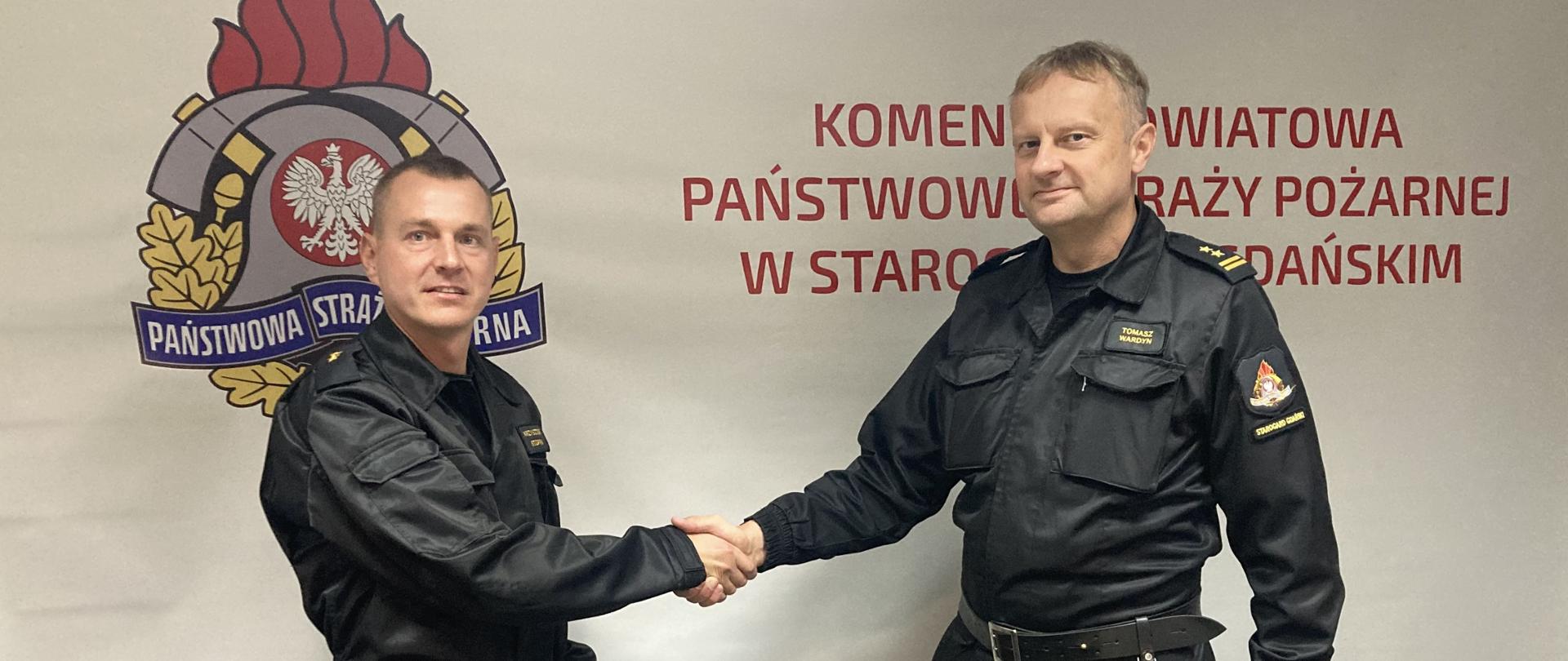 Zdjęcie przedstawia moment gratulacji, komendant powiatowy gratuluje nagrodzonemu funkcjonariuszowi, w tle logo PSP