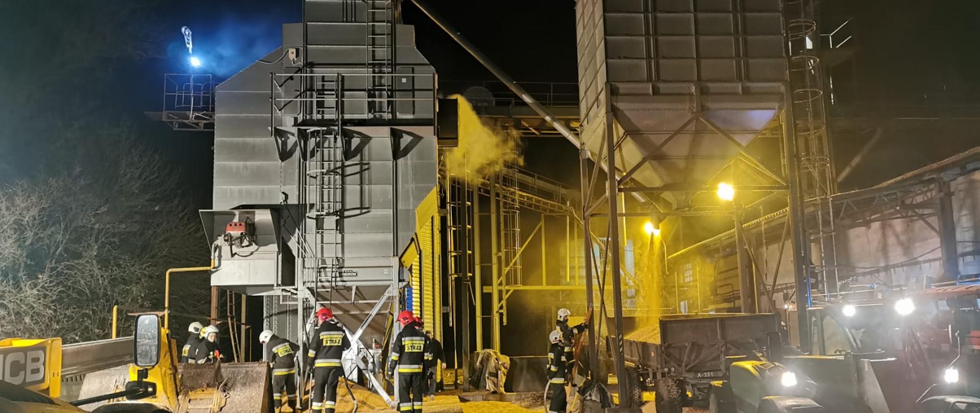 Pożar suszarni Piechanin. Na zdjęciu widać strażaków działających podczas pożaru suszarni kukurydzy. Widać suszarnie i zbiorniki. Kukurydza wysypywana jest na przyczepy.