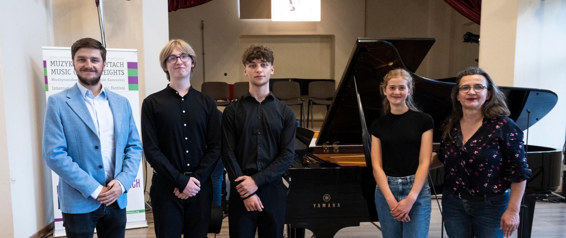 Na zdjęciu stoi pięć osób w sali koncertowej. W tle znajduje się fortepian marki Yamaha oraz kilka krzeseł ustawionych na podwyższeniu. Od lewej strony: Pierwsza osoba to mężczyzna w jasnoniebieskiej marynarce, białej koszuli i ciemnych spodniach.
Druga osoba to młody mężczyzna w okularach, ubrany całkowicie na czarno. Trzecia osoba, również młody mężczyzna, ubrany jest w czarną koszulę i czarne spodnie, z brązowymi butami. Czwarta osoba to młoda kobieta w czarnym T-shircie i niebieskich dżinsach, w białych sportowych butach. Piąta osoba to kobieta w okularach, ciemnej bluzce w kwiatowy wzór i ciemnych spodniach. W tle, po lewej stronie, widoczny jest stojak z plakatem zawierającym napisy, m.in. „Muzyka na Szczytach” oraz informację o festiwalu muzyki kameralnej. Na podłodze, w prawym dolnym rogu zdjęcia, znajduje się kolorowe logo z tym samym napisem.
Wszyscy stoją obok siebie i uśmiechają się w stronę aparatu.